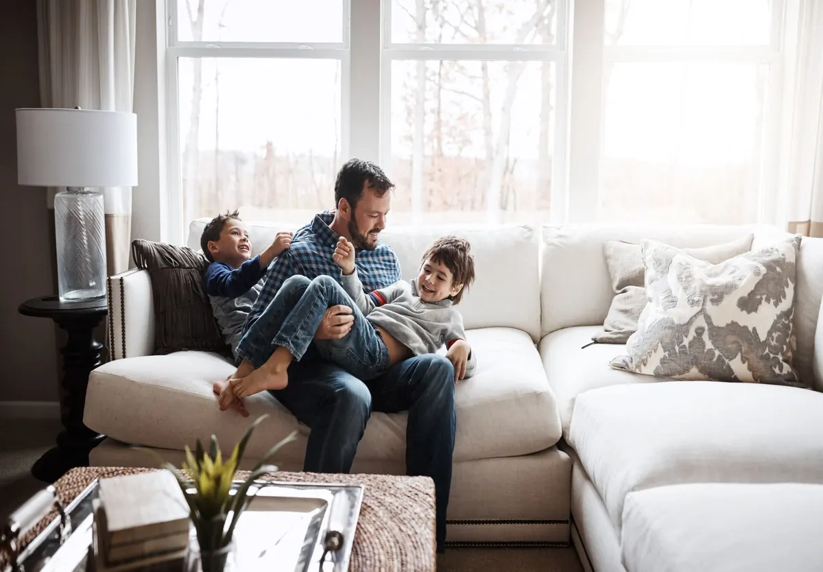 A father playing with his two sons on a white sofa in a bright living room