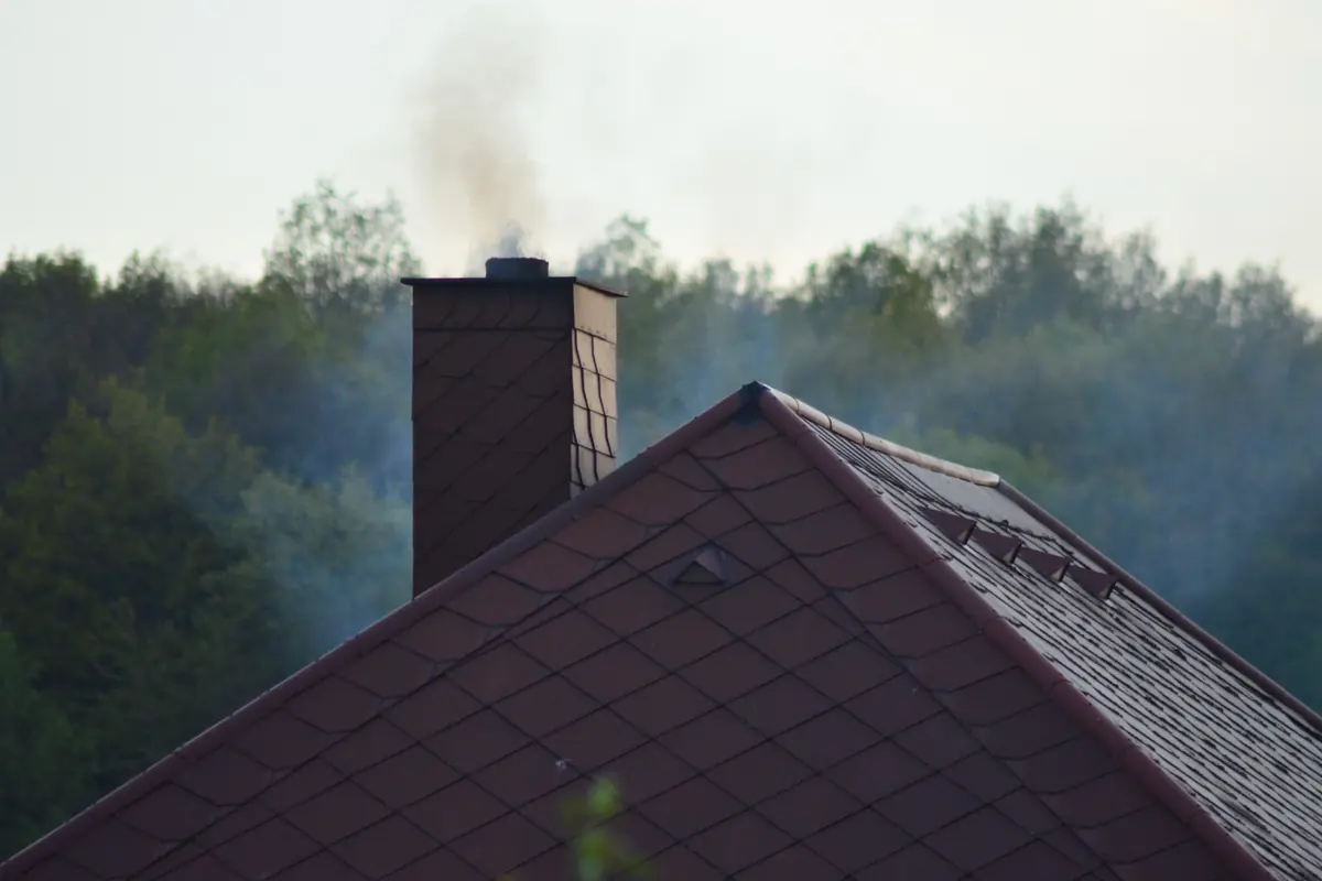 Smoke rising from a brick chimney on a dark roof with trees behind