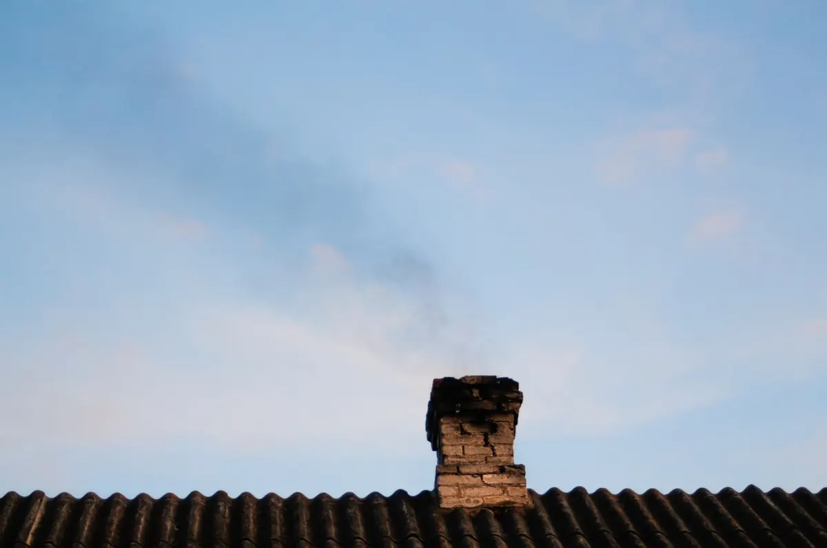 A weathered brick chimney with crumbling mortar on a tile roof under a blue sky